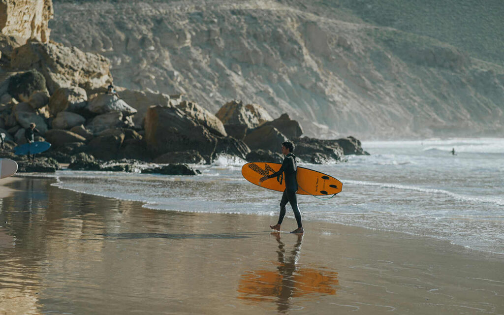 a guy with a surfboard walking on a beach in imsouane bay