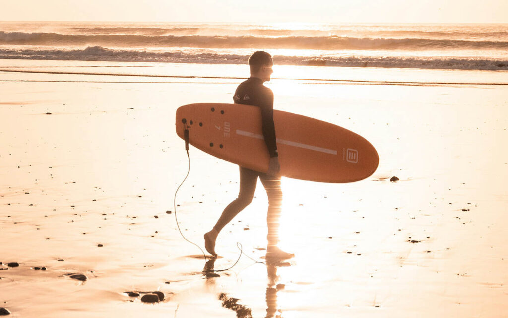 surfer walking with a softtop surfboard on the beach at sunset