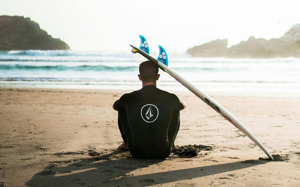 surfer sitting on the beach with his shortboard looking at the ocean